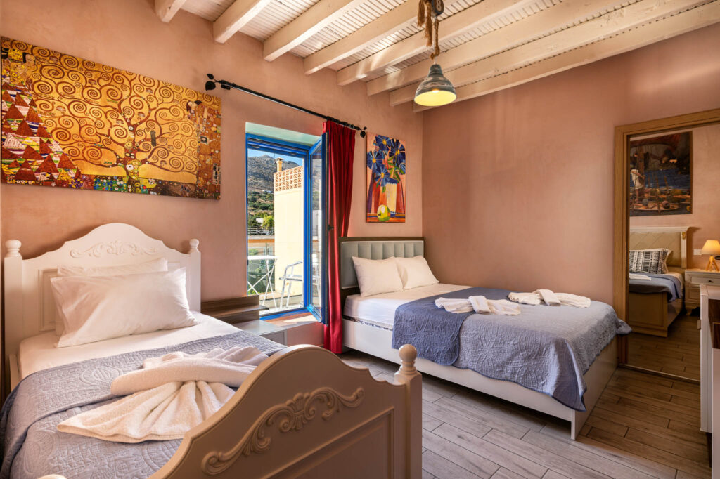 Bedroom at a holiday home in Zakros with 2 beds, colorful paintings and red curtains to a small balcony with mountain view.