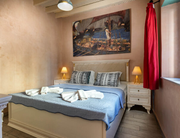 A king size bed with blue-white sheets and pillows in a room with a picture on the wall and red curtains at the blue window